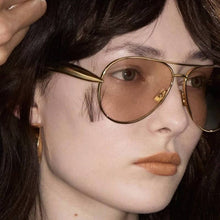 Load image into Gallery viewer, Close-up of a person wearing gold-framed sunglasses with a blurred background
